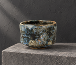 Ambientazione creata con IA : Chawan blu grigio con zone scure, su un piedistallo.