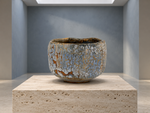 AI-generated setting : Pale blue chawan with an ochre mosaic, on a travertine plinth.