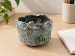 Ambientazione creata con IA : Chawan verde grigio con reticolo nero, su una scrivania con piccoli oggetti.
